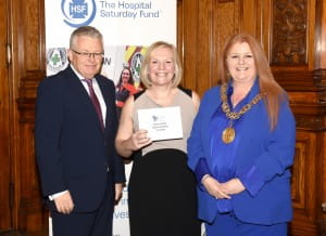 3 individuals stood, the middle person is posing with a cheque in hand. 