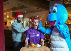 SBH Scotland CEO is sandwiched between SBH Scotland's dinosaur mascot and Street Burger Manager Tim. He is having a burger bun with relish smushed on each side of his face and looking shocked