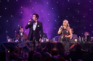 Nicola Cassells and Colin Bryce are stood on a dramatically lit stage singing. They are dressed in tartan finery. 