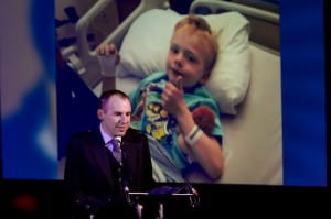 Sean Carr is standing at a podium on a dramatically lit stage with an image of his child on a screen behind. He is talking to an audience at a Question of Burns Supper.