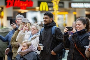 group of shoppers have gathered together in a shopping centre to watch a performance. Some are filming the performance and others smiling. 