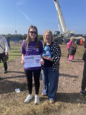 2 ladies stood next to each other. One holding a toy dinosaur and the other a certificate. 