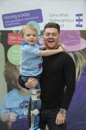 Reece Donnelly stood smiling to camera. Reece is lifting a young boy with charity t-shirt up. Both are in front of a charity banner.
