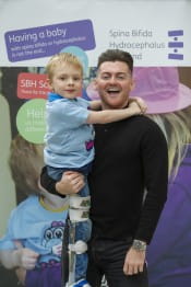 Reece Donnelly stood smiling to camera. Reece is lifting a young boy with charity t-shirt up. Both are in front of a charity banner.